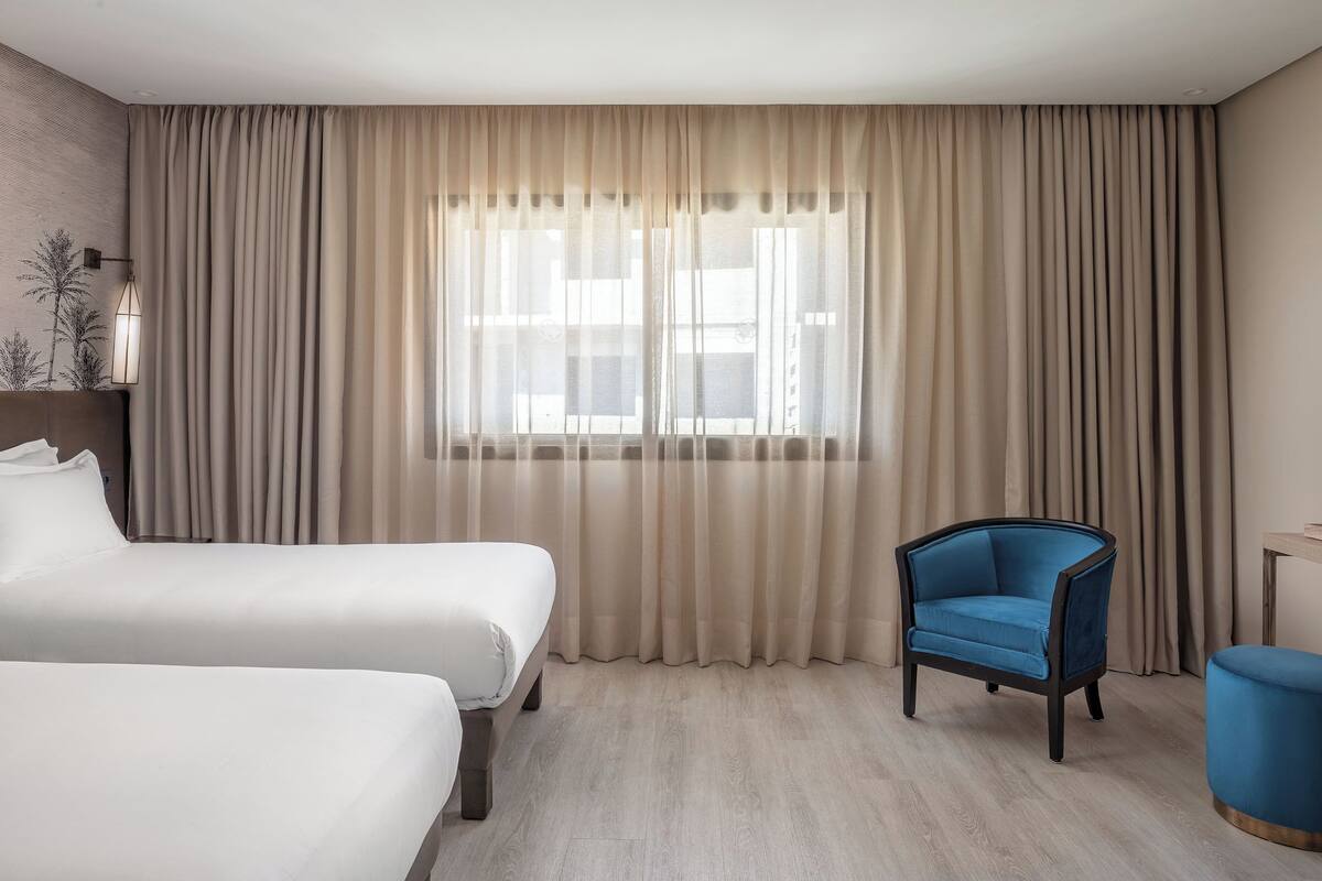Habitación de hotel con dos camas, un sillón azul y una mesita.
