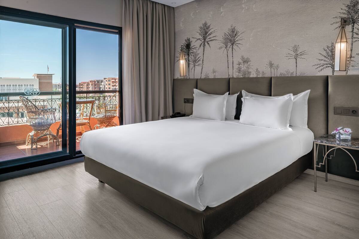 Una cama grande con cabecera, una mesita de noche, una lámpara y vistas de la ciudad a través de amplias ventanas.