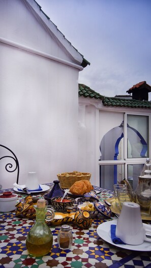 A table set with various dishes and a bottle, a patterned floor, and a white building with a green roof. - Hotel Casa Miguel (Chefchaouen)