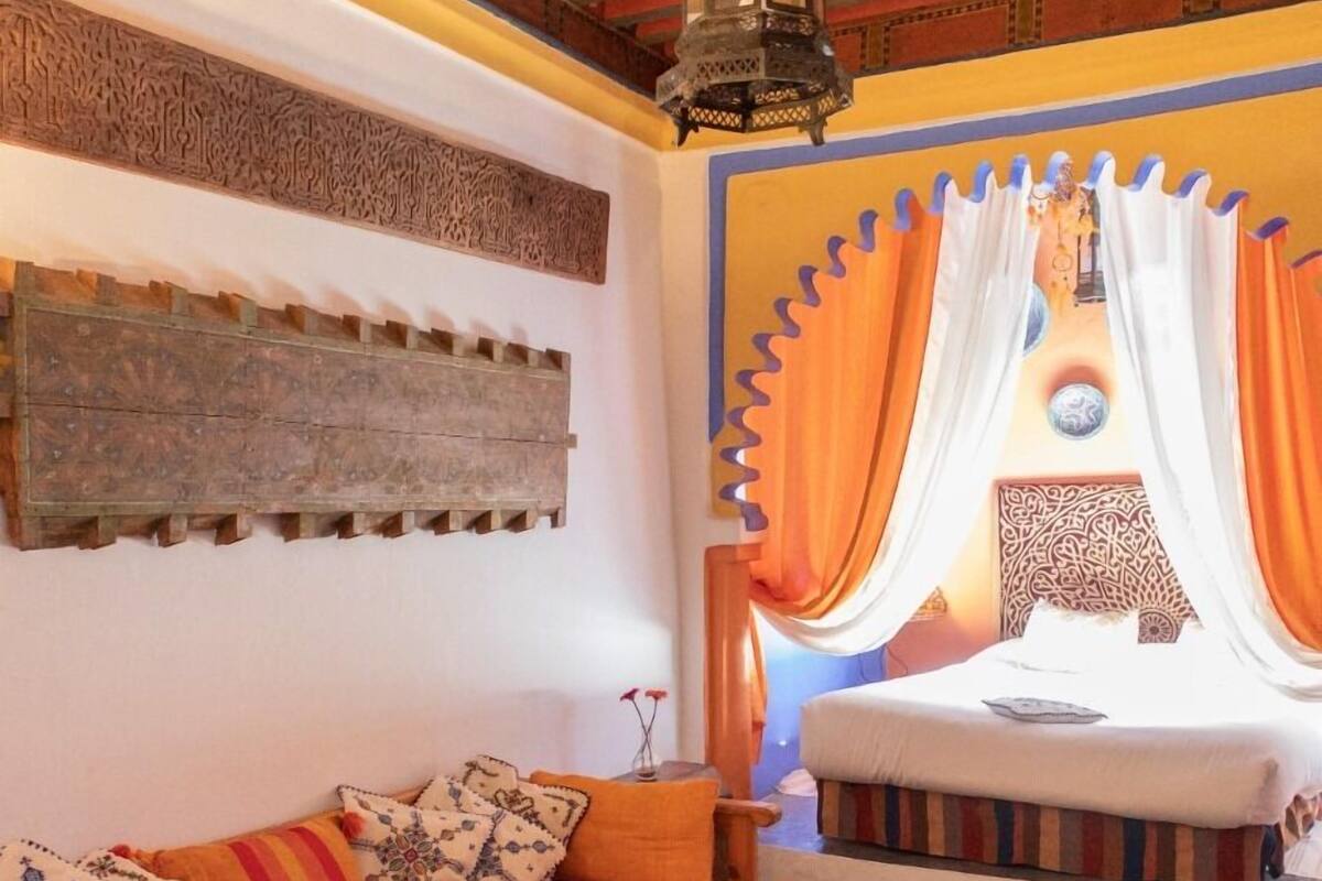 A cozy room with a wooden sofa, a bed with orange curtains, and a patterned rug.
