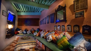 Un cuarto acogedor con un largo banco decorado con almohadones de colores, un televisor sobre un mueble bajo y cuadros enmarcados en la pared.