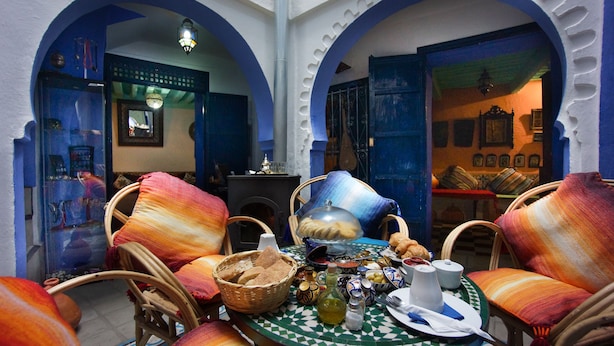 Un interior tradicional marroquí con un comedor que incluye sillas de mimbre, una mesa dispuesta con pan, aceitunas y una botella de aceite de oliva.