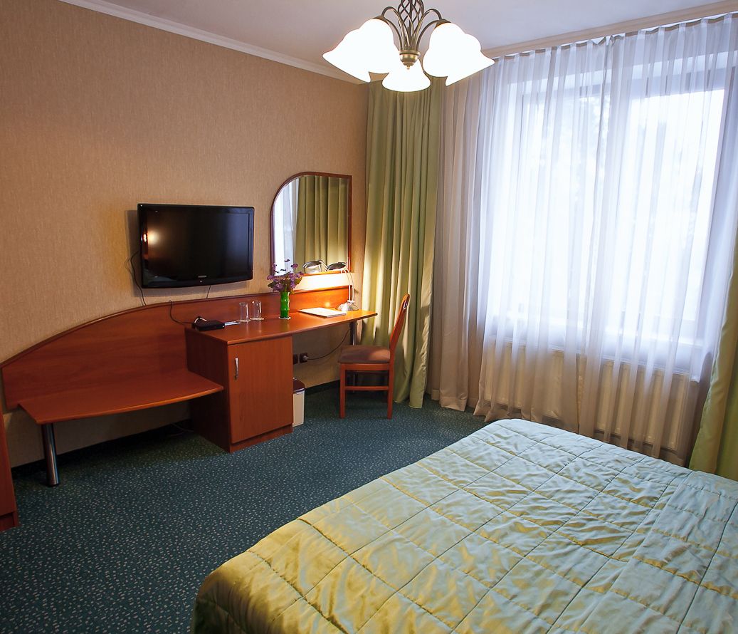 Ein Hotelzimmer mit Bett, Schreibtisch, Stuhl, Fernseher und einem Fenster mit Vorhängen.