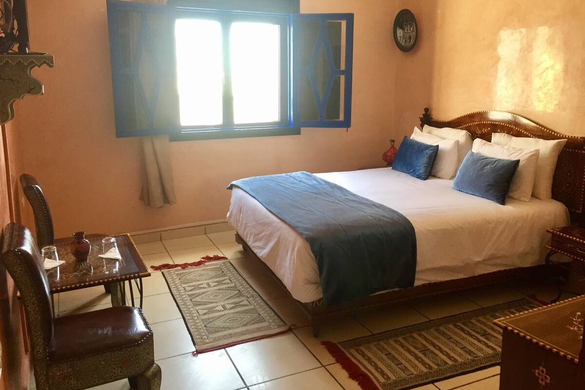 A bedroom with a bed, a table with chairs, a window, and a wall clock.