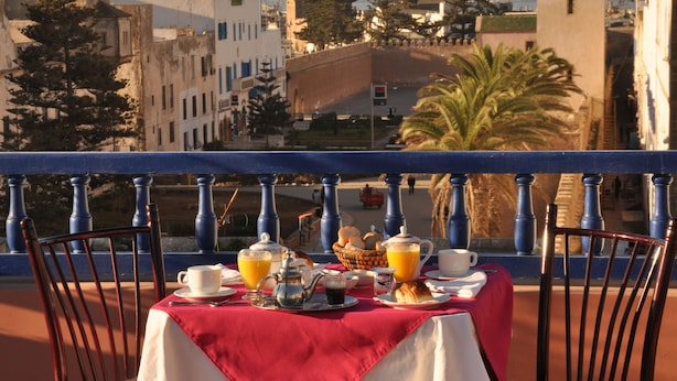 A table set with breakfast items, including a basket of bread, a teapot, cups, and orange juice, on a balcony overlooking a cityscape with buildings and a clock tower. - Essaouira Wind Palace (Essaouira)