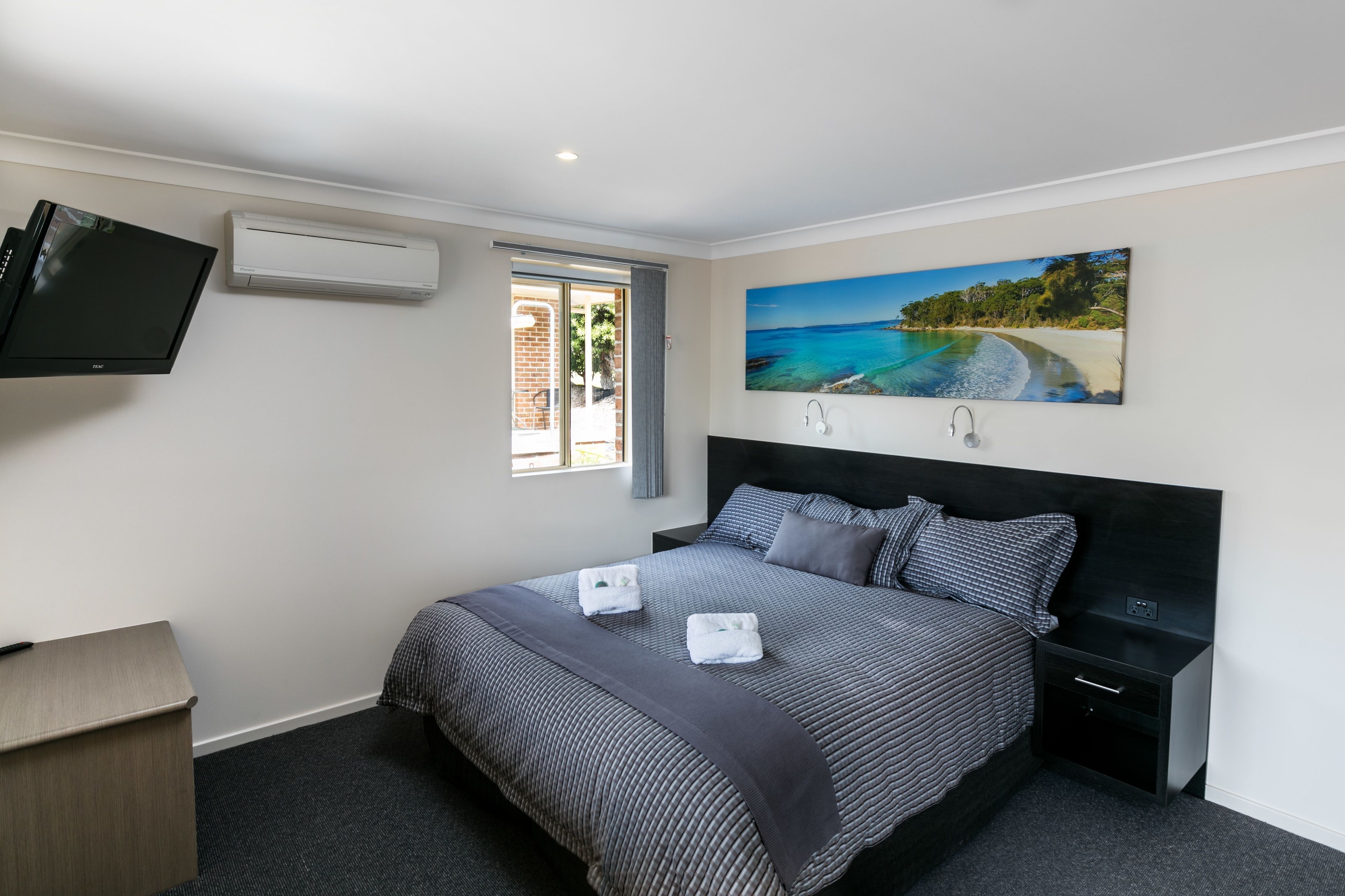 Un dormitorio con cama, televisor, aire acondicionado, un cuadro de una playa y una ventana.