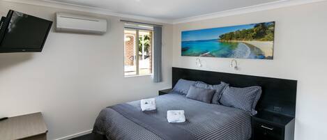 A bedroom with a bed, a TV, an air conditioner, a painting of a beach, and a window.