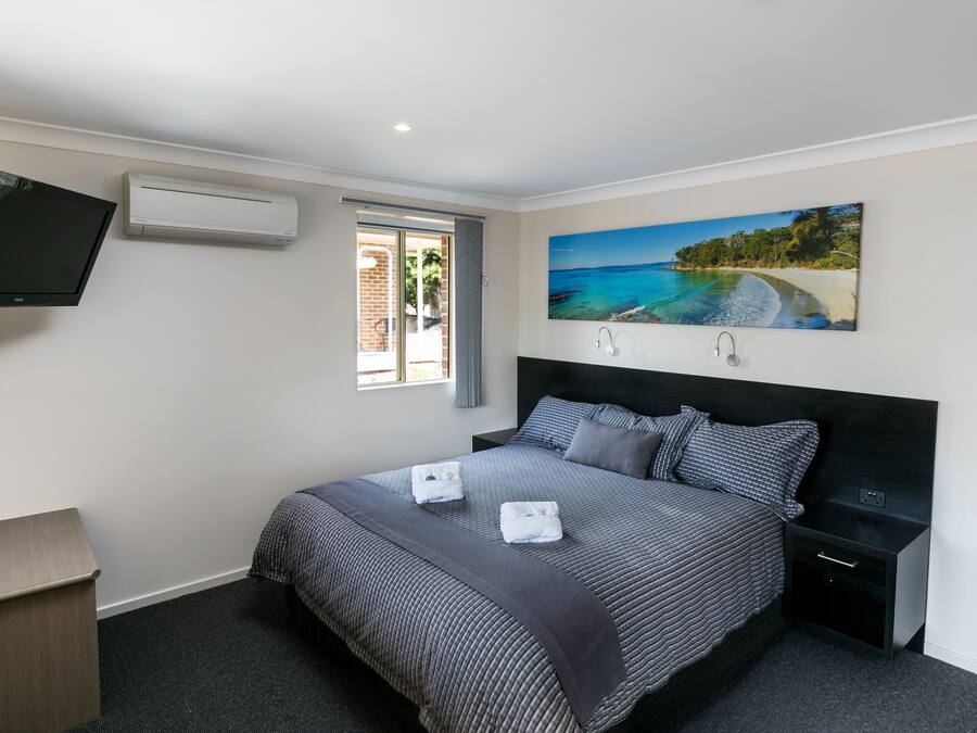 Un dormitorio con cama, televisor, aire acondicionado, un cuadro de una playa y una ventana.