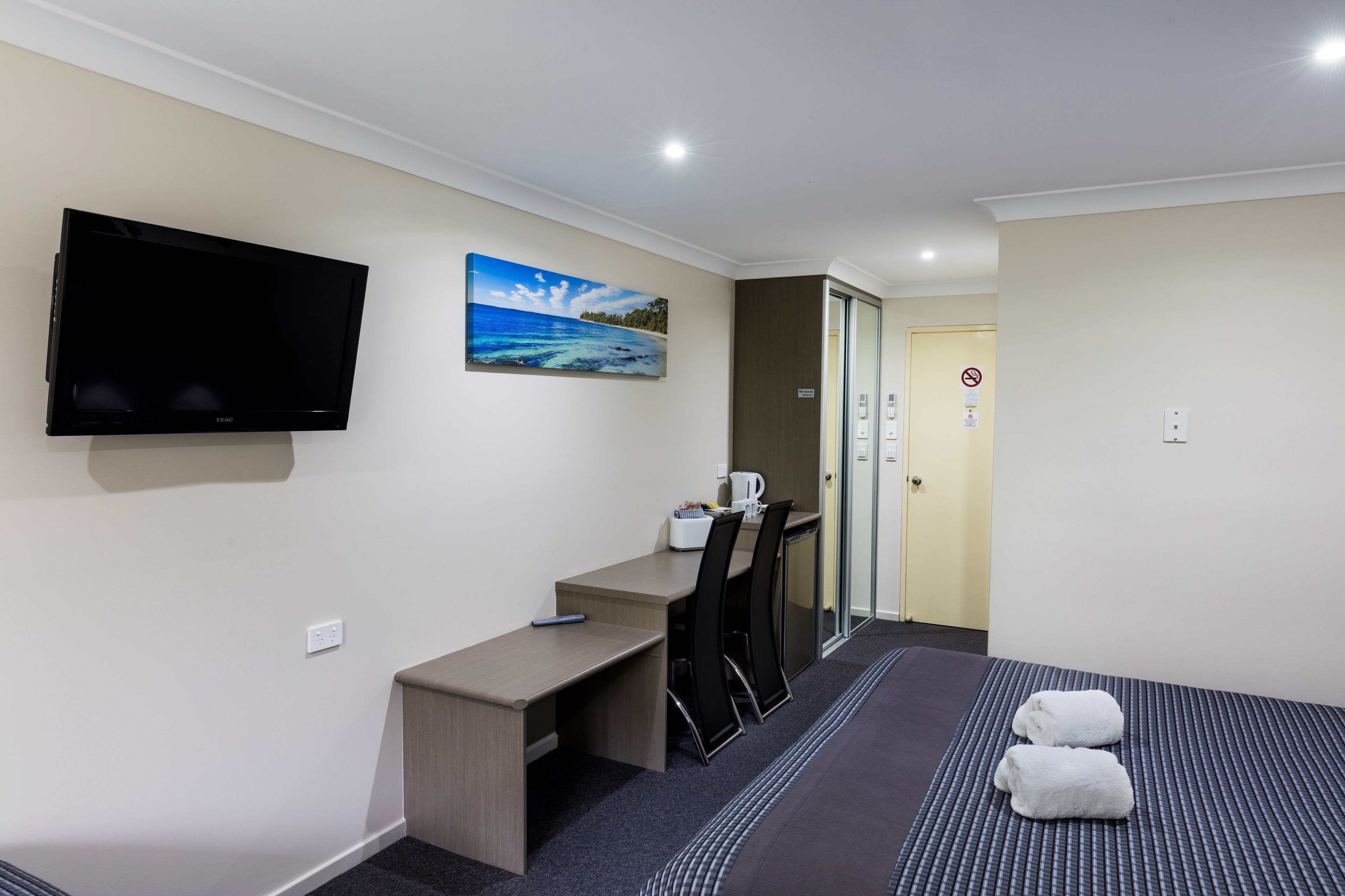 Een hotelkamer met een bed, een bureau, een stoel, een flatscreen-tv en een schilderij van een strandgezicht.