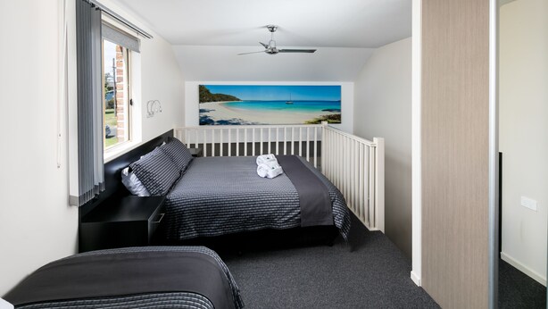 A bedroom with a bed, a view of a beach, a ceiling fan, and a window with curtains. - Dolphin Shores (Vincentia)