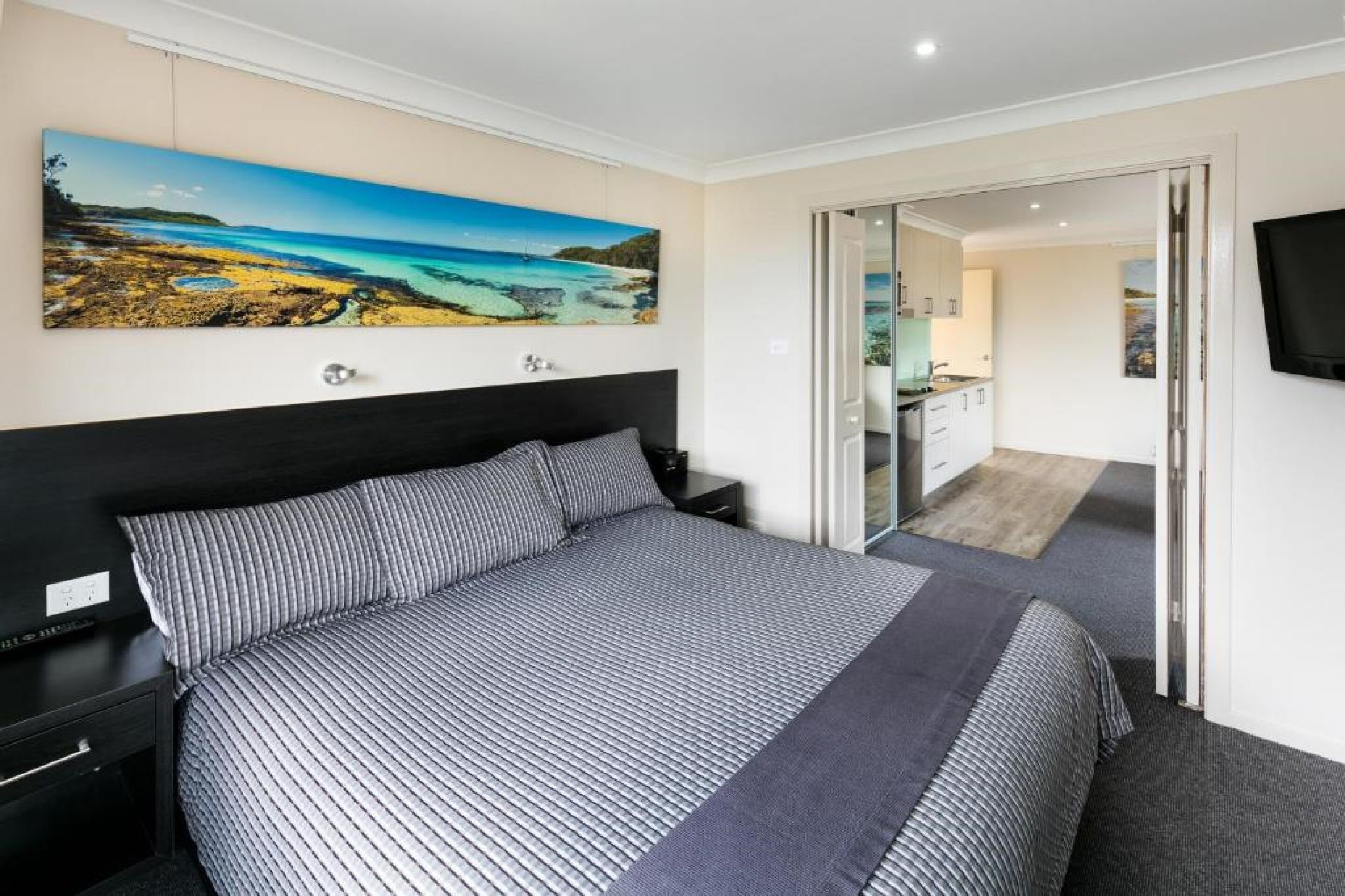 A bedroom with a large bed, a nightstand, a painting of a coastal landscape, a TV mounted on the wall, and a sliding door leading to a bathroom.