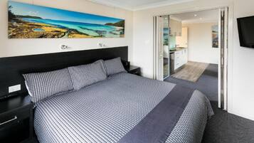 A bedroom with a large bed, a nightstand, a painting of a coastal landscape, a TV mounted on the wall, and a sliding door leading to a bathroom.