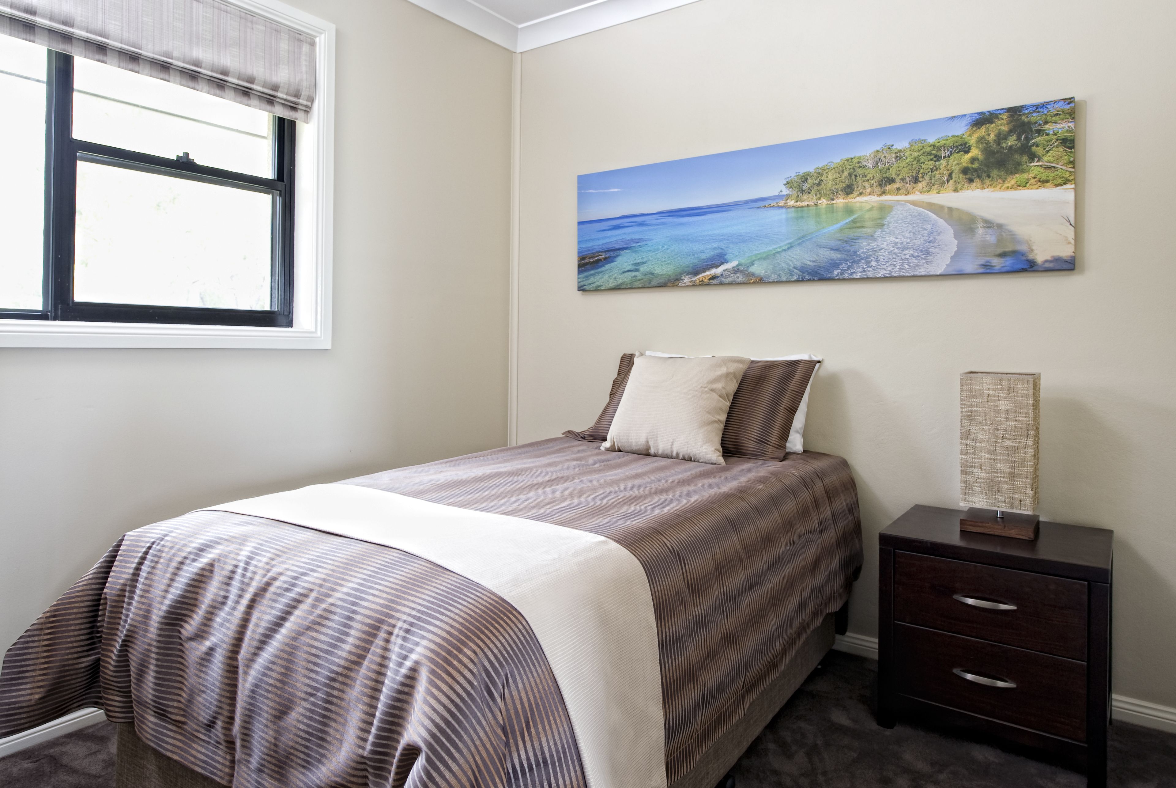 Un dormitorio con una cama, una mesita de noche, una ventana y un cuadro con una escena de playa en la pared.