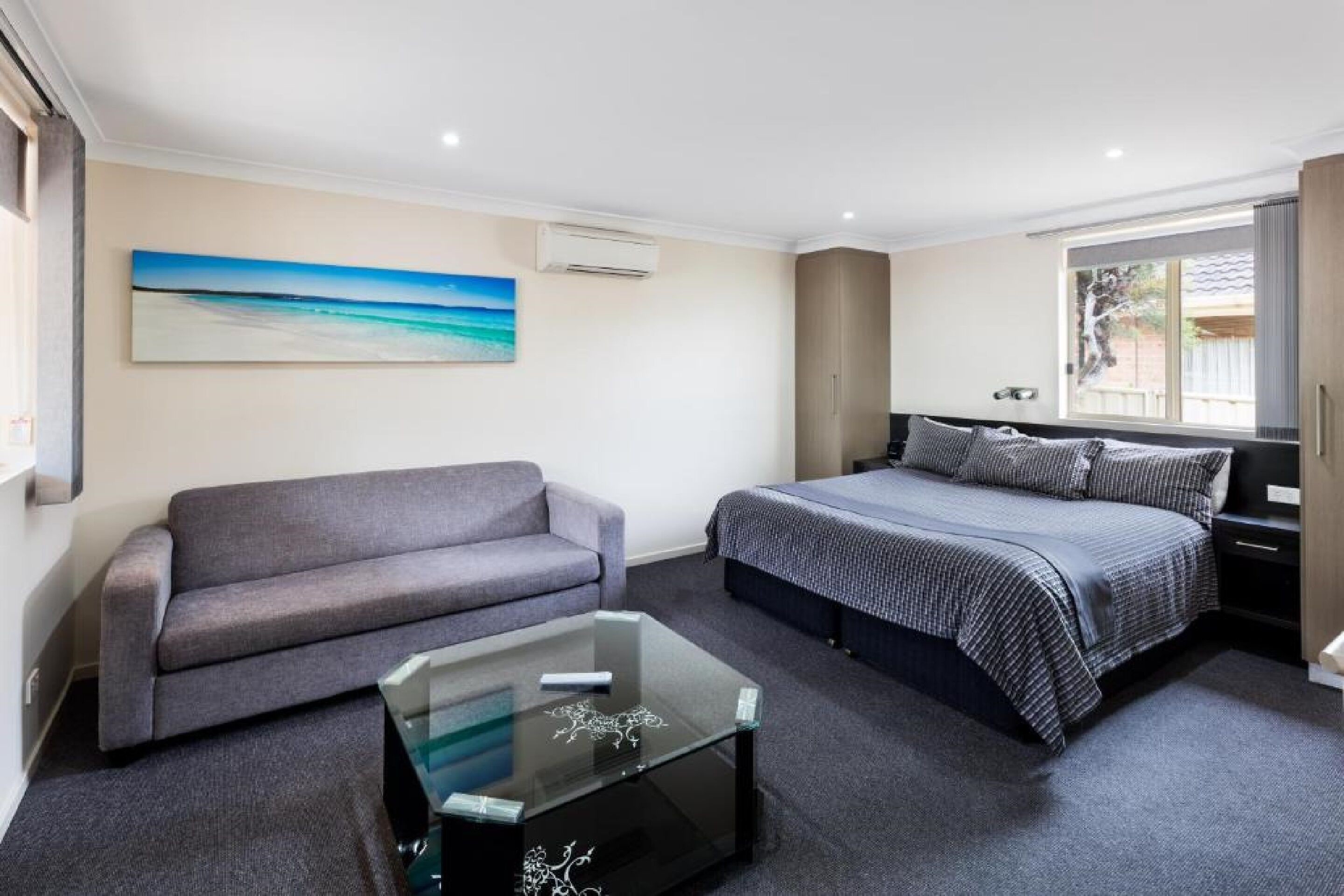 A bedroom with a bed, a sofa, a glass coffee table, and a painting on the wall.