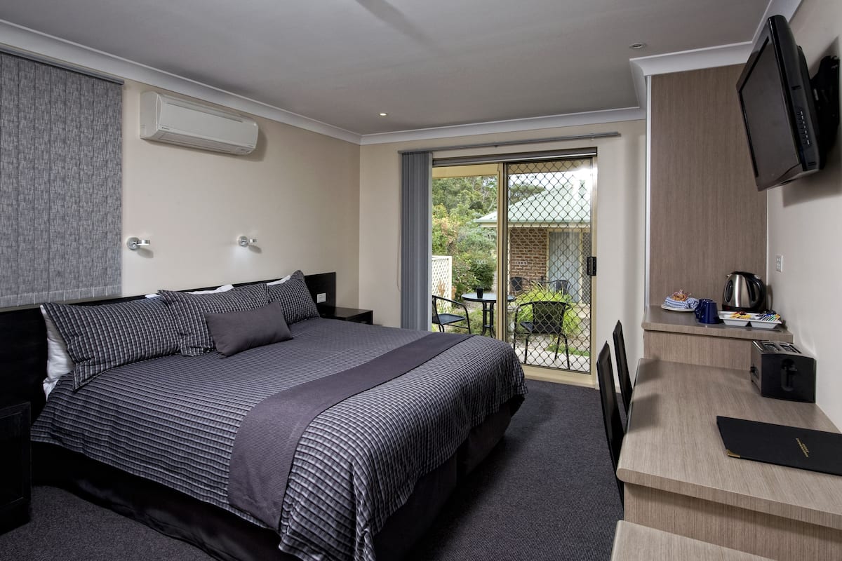 A modern hotel room with a large bed, a desk with a flat-screen TV, a coffee maker, and a sliding glass door leading to a balcony.