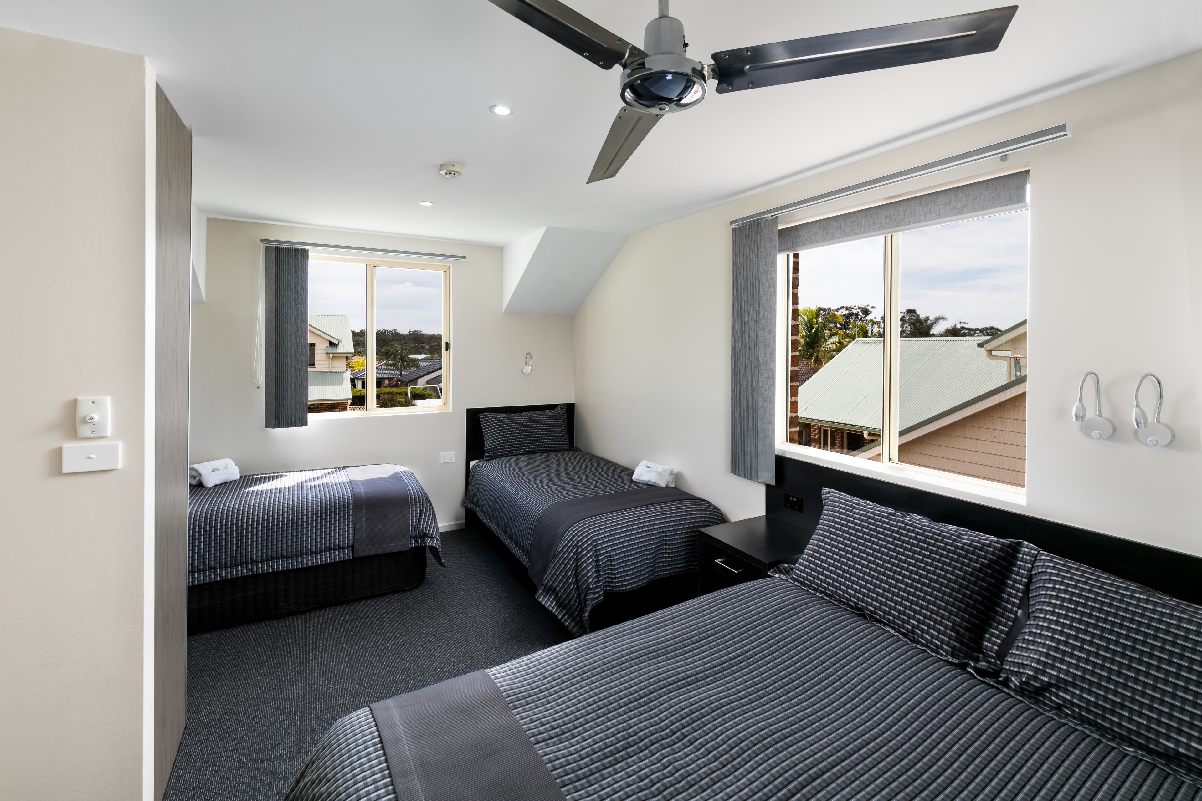 Una habitación de hotel moderna con dos camas individuales, un ventilador de techo, un ventanal con vistas y un pequeño escritorio con una silla.