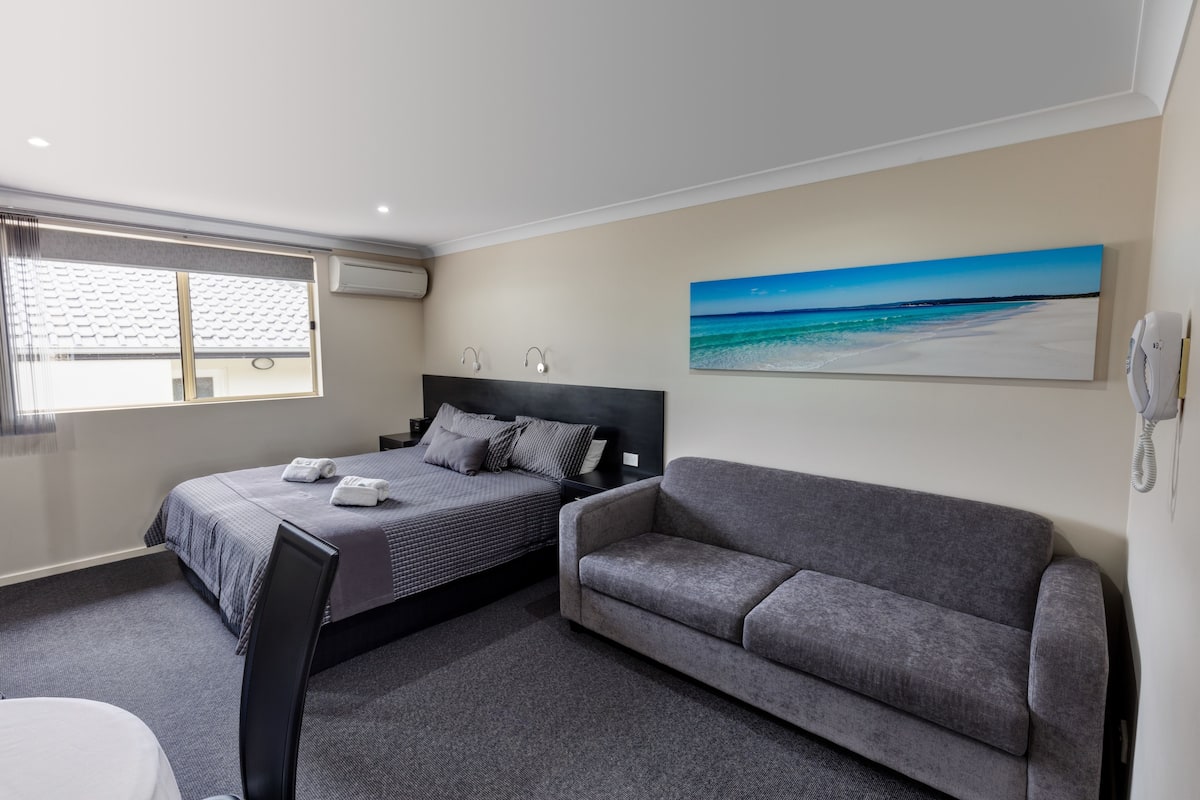 A hotel room with a bed, a gray sofa, a painting of a beach, and a phone on the wall.