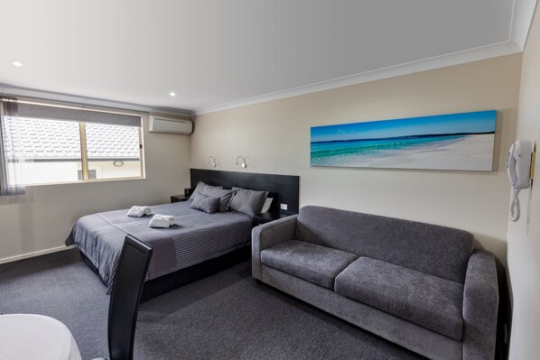 A hotel room with a bed, a gray sofa, a painting of a beach, and a phone on the wall.