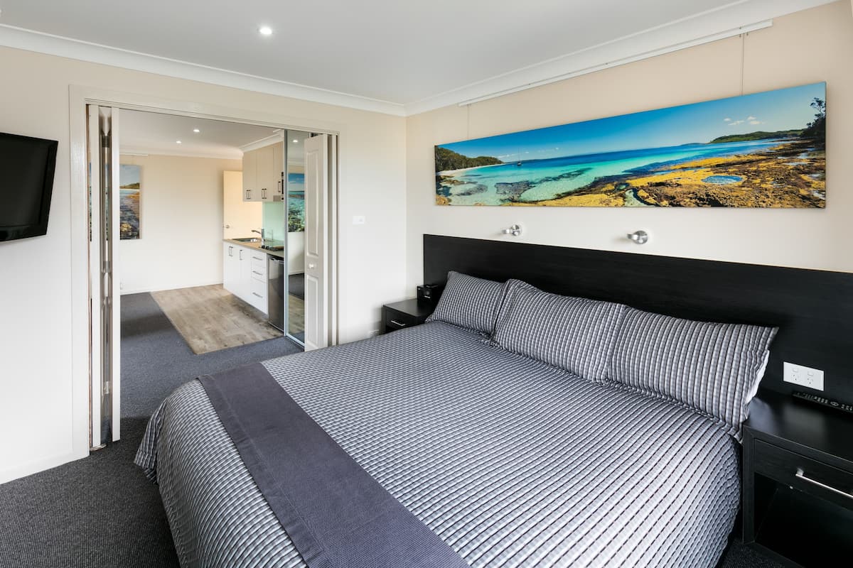 A bedroom with a large bed, a painting of a beach scene, a TV mounted on the wall, and a door leading to a bathroom.