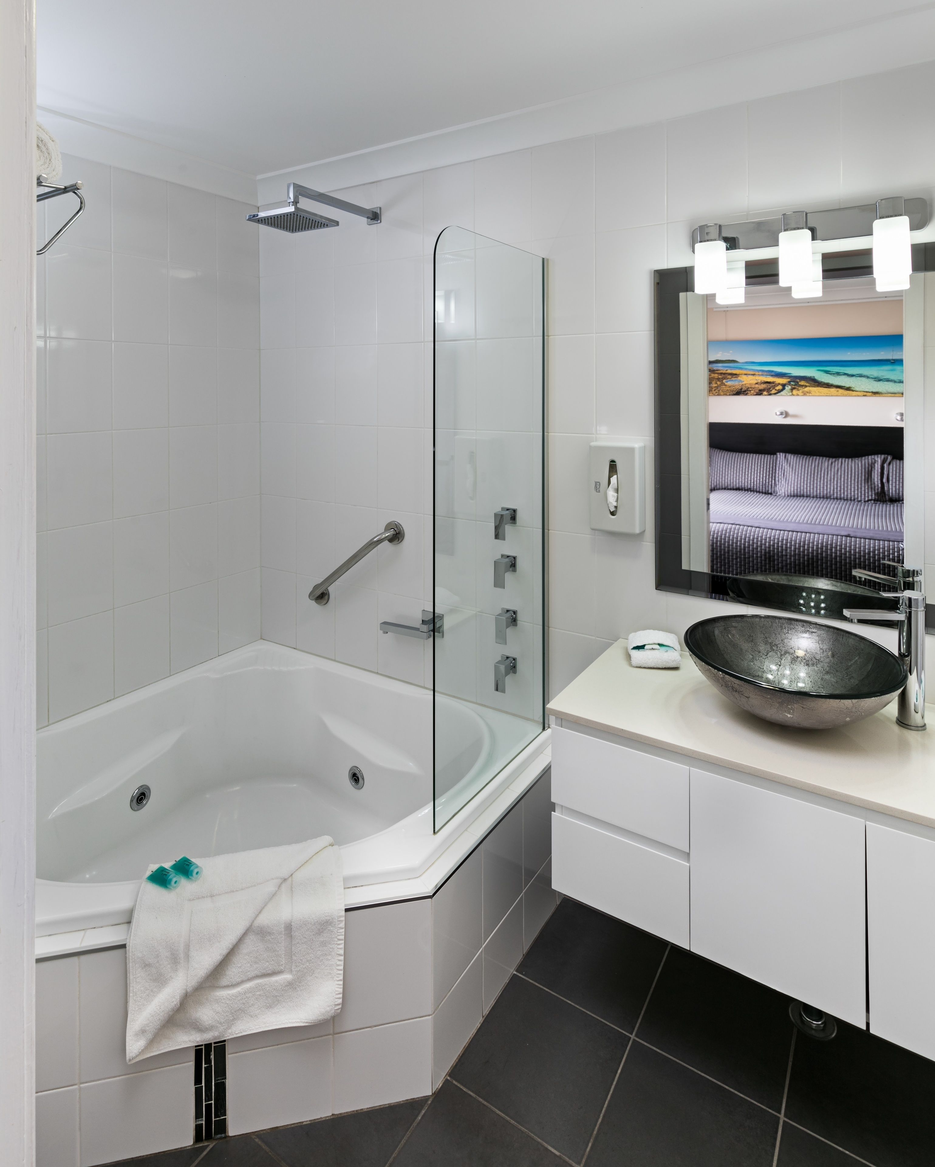 A modern bathroom with a glass-enclosed shower, a white bathtub, a large mirror with a painting, and a black basin sink.