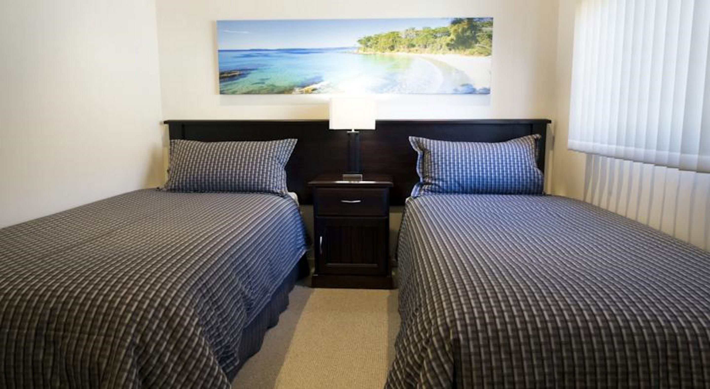 A hotel room with two single beds, a nightstand with a lamp, and a painting of a coastal scene.