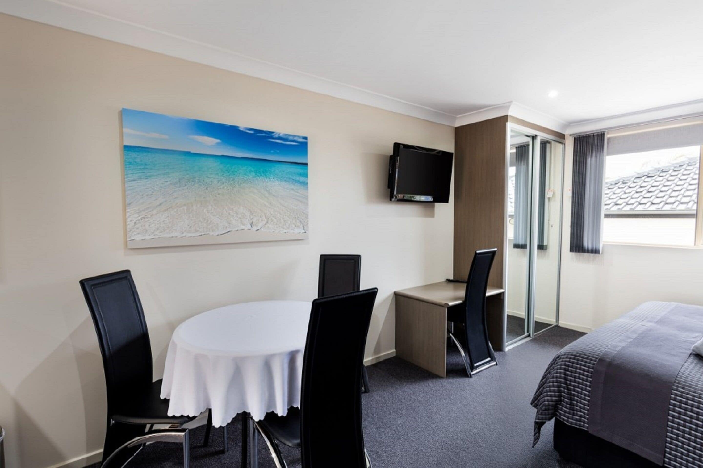 Habitación de hotel con una mesa redonda, dos sillas, una cama, un televisor y un cuadro de una playa.