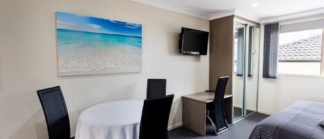 A hotel room with a round table, two chairs, a bed, a TV, and a painting of a beach.