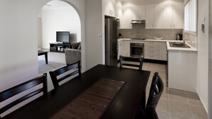 A modern kitchen with white cabinets, a stainless steel refrigerator, and a built-in oven. There is a dining area with a dark wooden table and chairs. - Dolphin Shores (Vincentia)