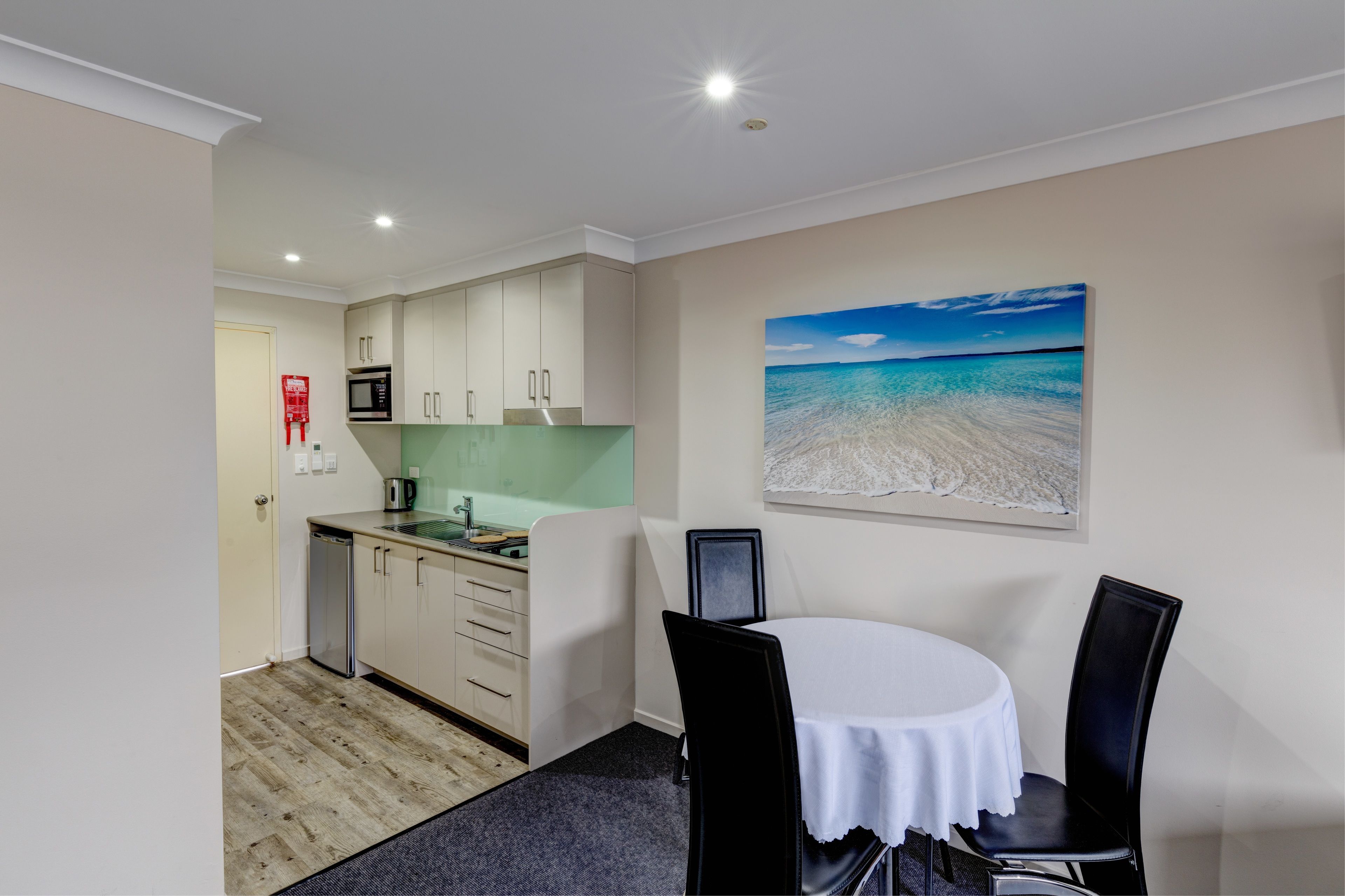 A compact kitchen with a microwave, sink, and cabinets, a dining table with chairs, and a beach scene painting on the wall.