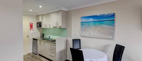 A compact kitchen with a microwave, sink, and cabinets, a dining table with chairs, and a beach scene painting on the wall.