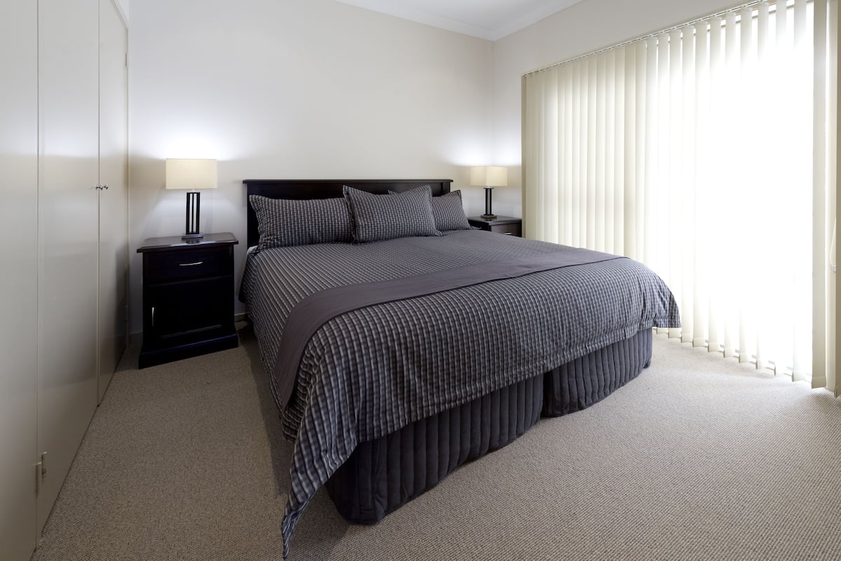 A neatly arranged bedroom with a large bed, two bedside tables with lamps, a wardrobe, and a window with blinds.