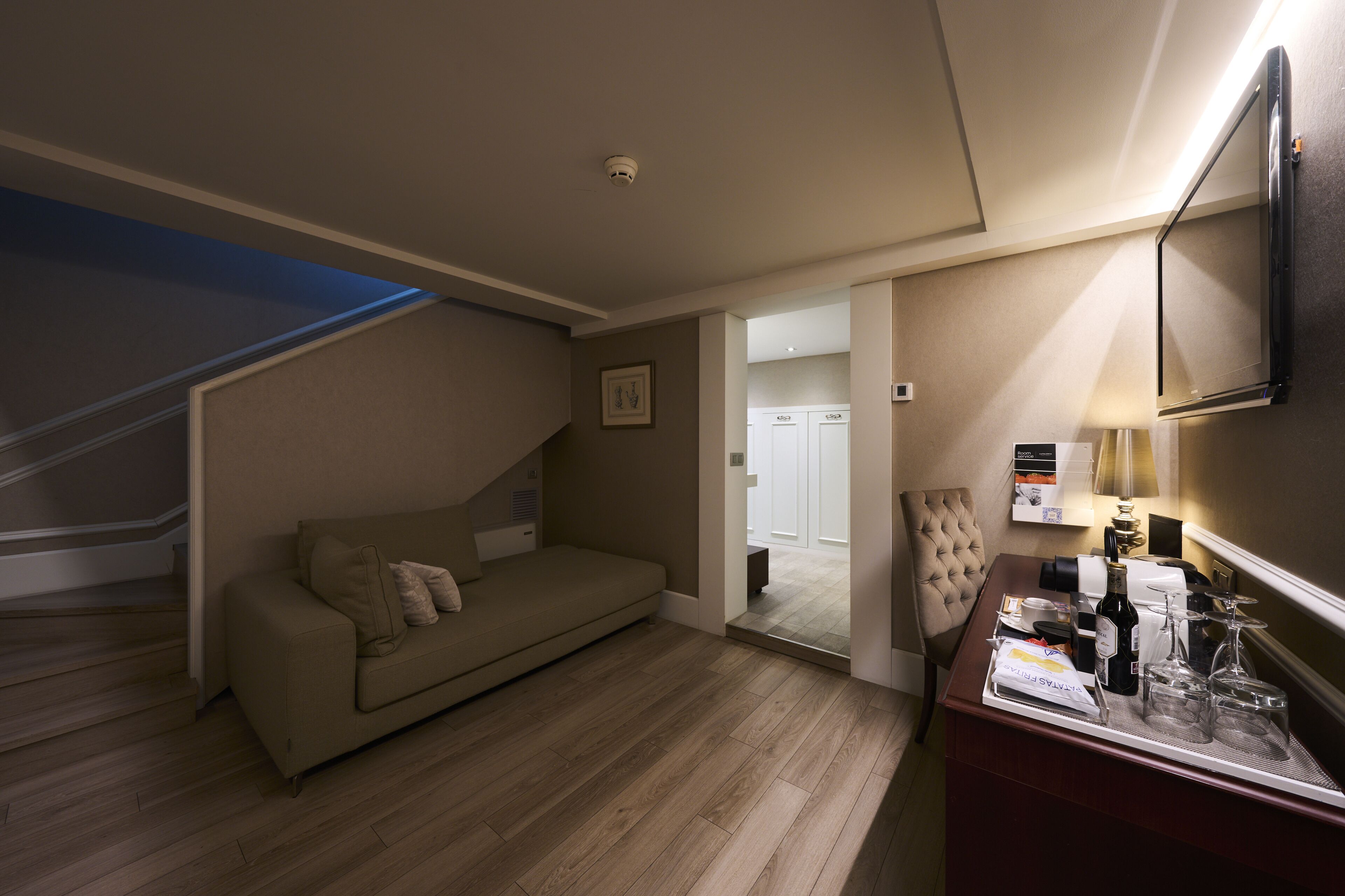 A hotel room with a sofa, a desk with a tray of glasses and a bottle, and a staircase.