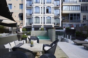 Een terras op het dak met buitenmeubilair, een zwembad en uitzicht op een meerhoog gebouw met balkons.