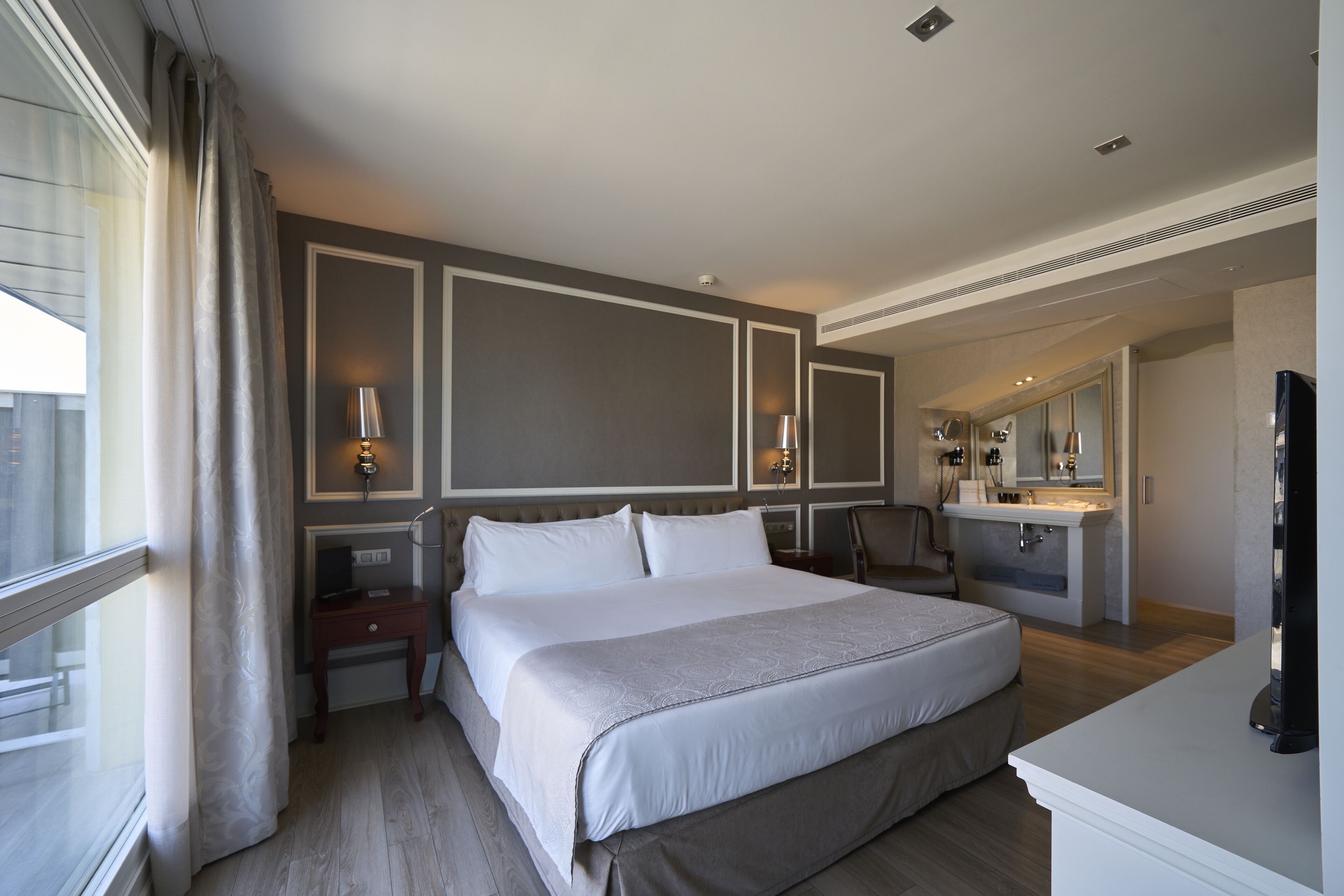 A hotel room with a large bed, bedside tables, a desk, a chair, a vanity with a mirror, and a television.