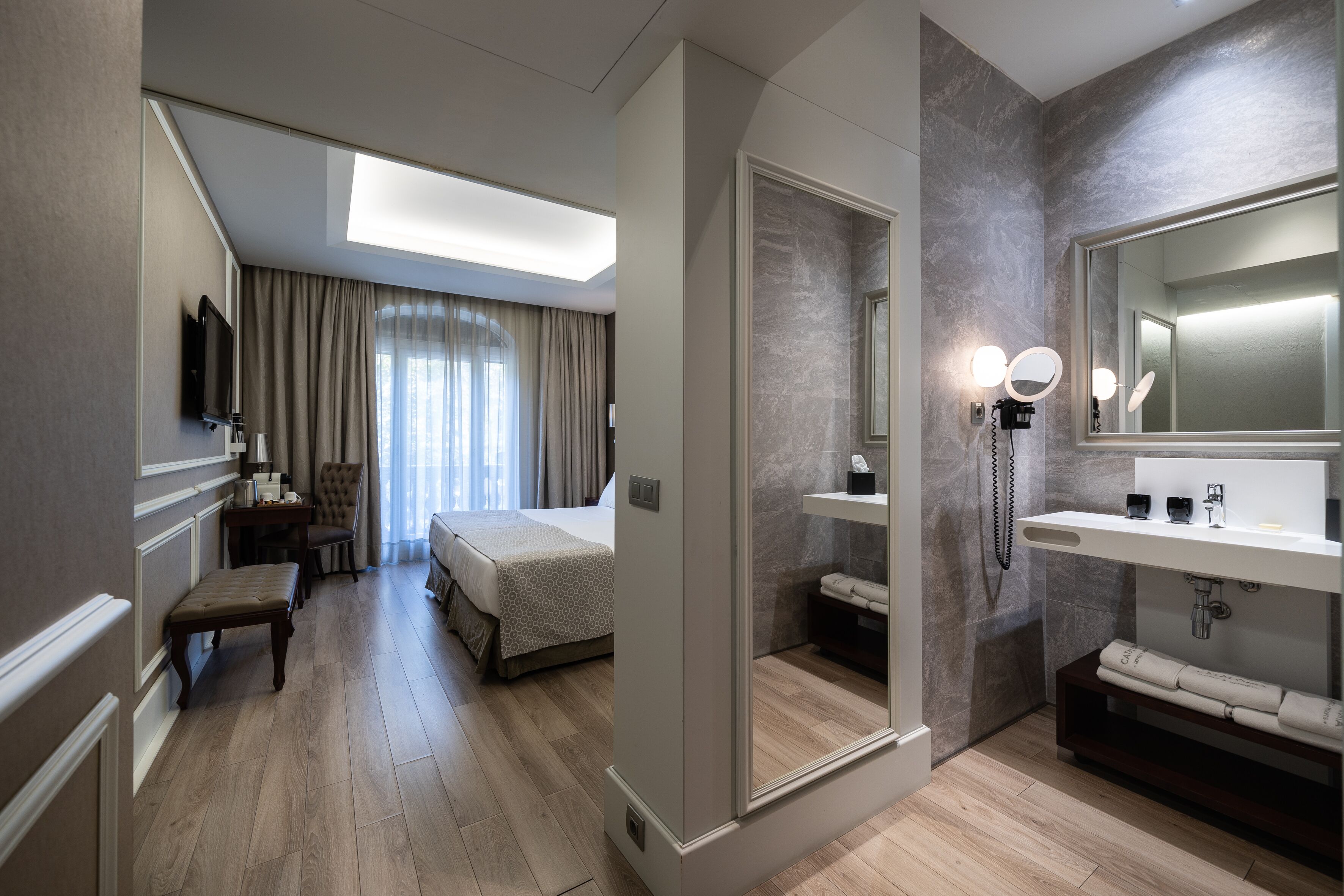 A hotel room with a bed, a desk, a chair, a bench, a bathroom with a mirror, sink, and towel rack.