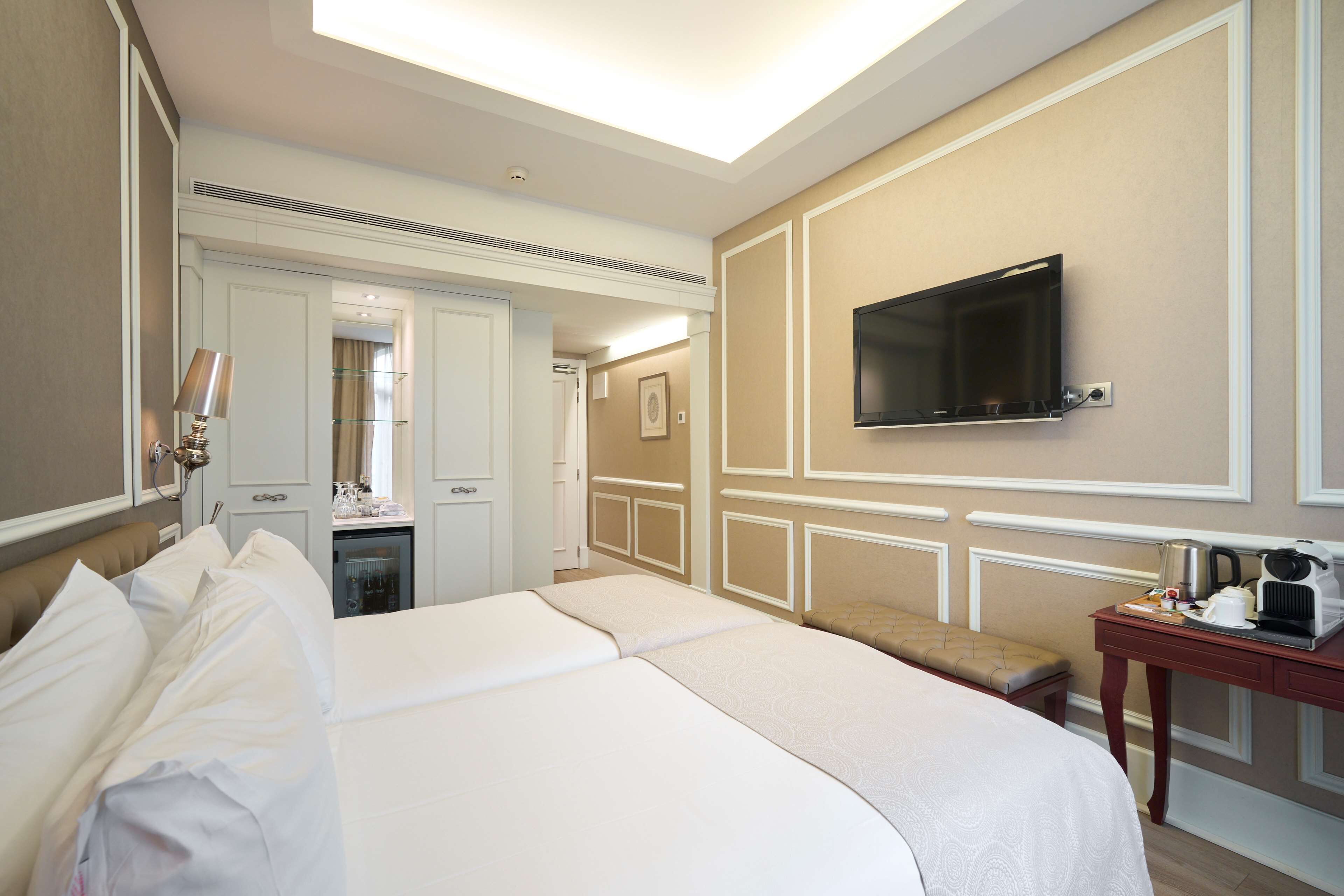 Una camera d'albergo con un letto grande, una TV fissata al muro, una piccola scrivania con un bollitore e un bagno visibile attraverso una porta aperta.