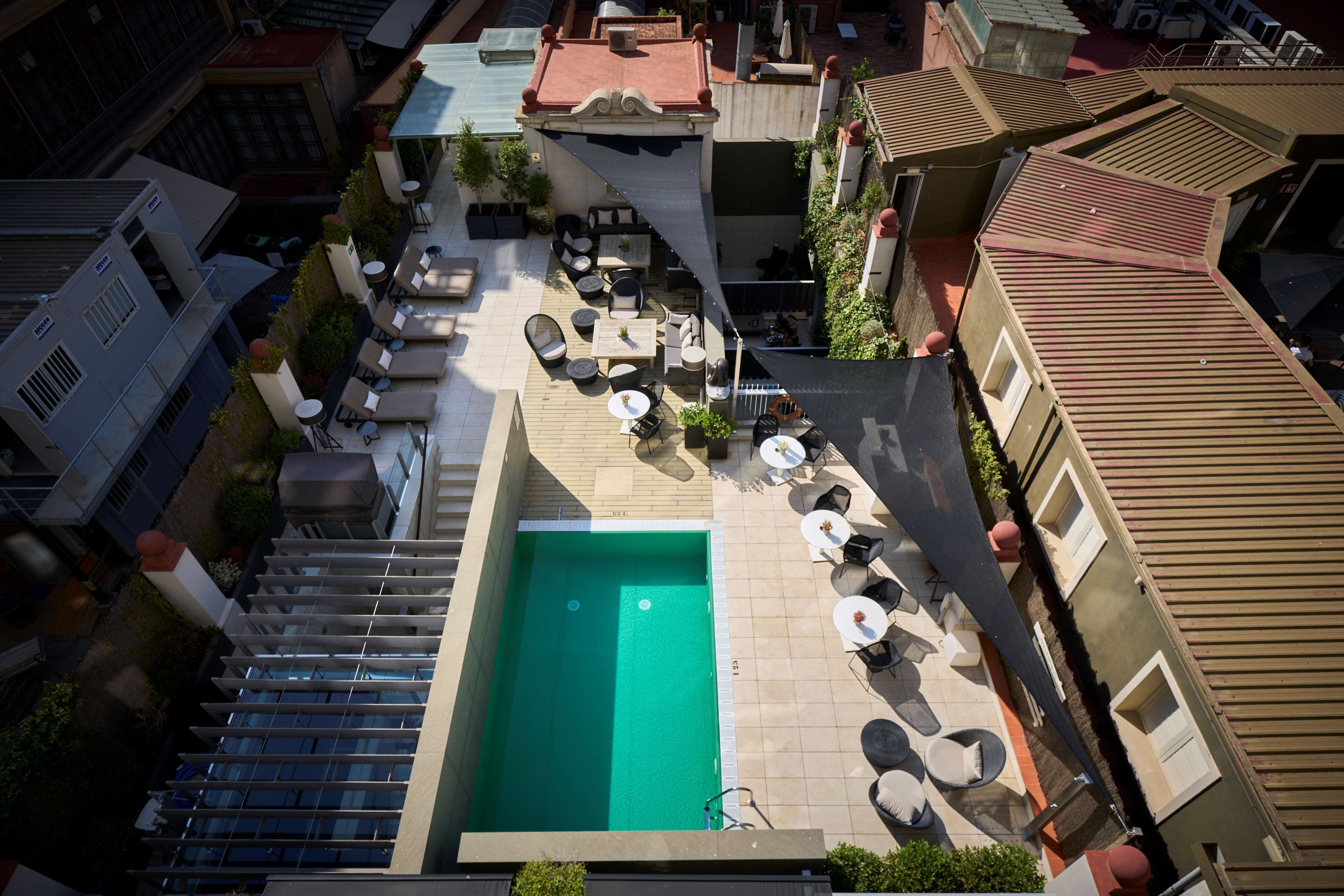 Vue aérienne d’un espace piscine sur le toit, avec plusieurs chaises longues et tables, le tout entouré d’immeubles.