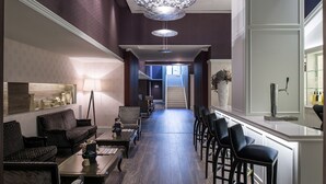 Un vestíbulo de hotel con un largo mostrador de recepción, área de descanso con sofás y sillones, una escalera que conduce a pisos superiores y una lámpara decorativa.