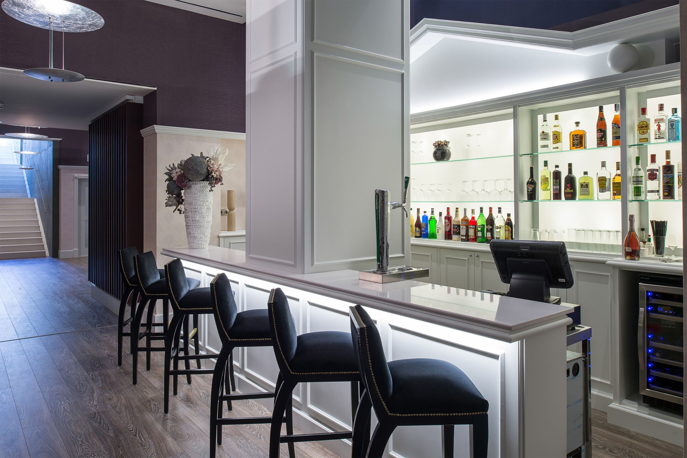 Una zona de bar con una barra alargada, taburetes negros y una vitrina de vidrio con varias botellas.