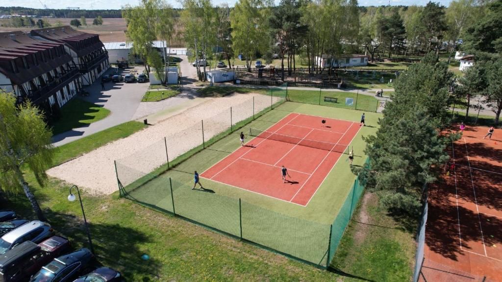 A tennis court surrounded by a fence, with a parking area and buildings in the background.
