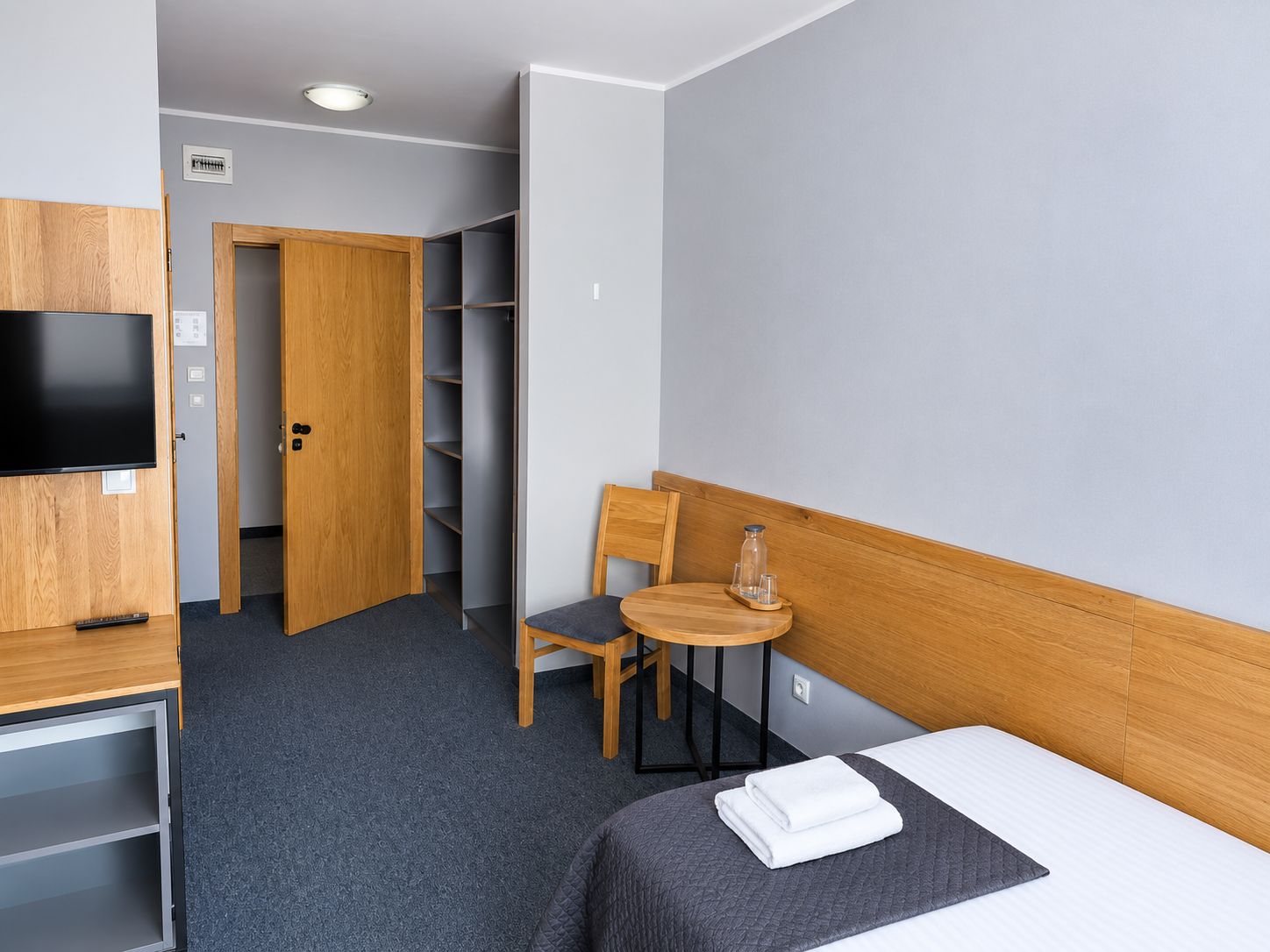 Habitación de hotel con cama, escritorio con televisor, silla, mesita con vaso y armario.