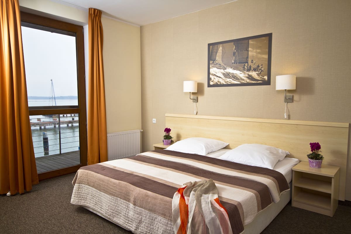Habitación de hotel con una cama, una mesita de noche, una lámpara, un jarrón con flores, una ventana con cortinas y un cuadro en la pared.