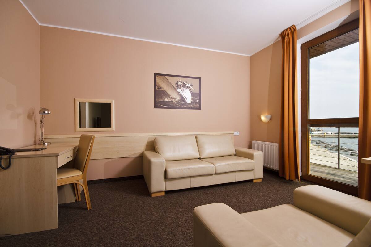 Habitación de hotel con sofá color beige, escritorio con lámpara, cuadro en la pared y vista al mar a través de un ventanal grande.
