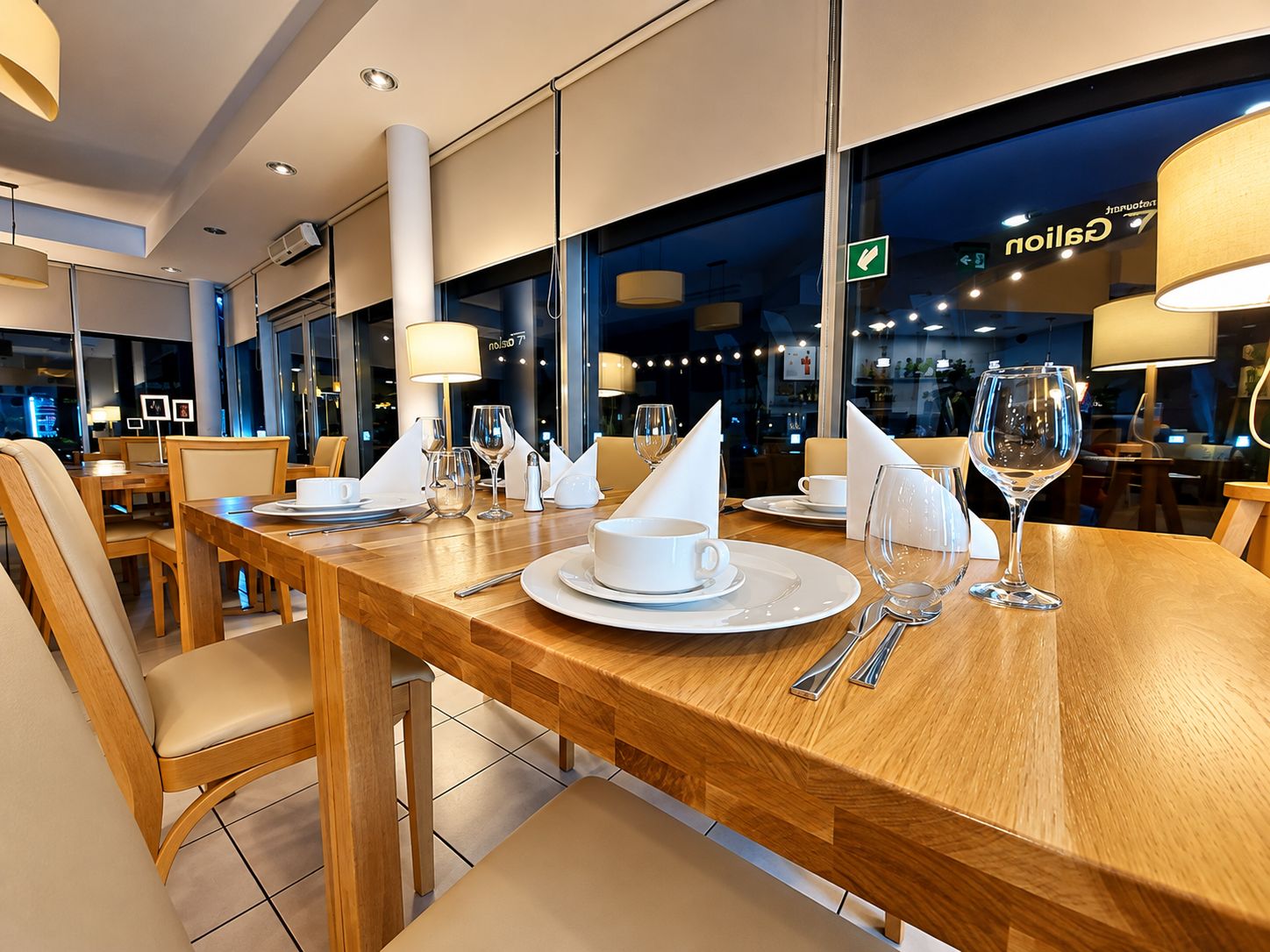 Un comedor bien iluminado con mesas de madera, vajilla blanca y vistas de la ciudad a través de amplias ventanas.