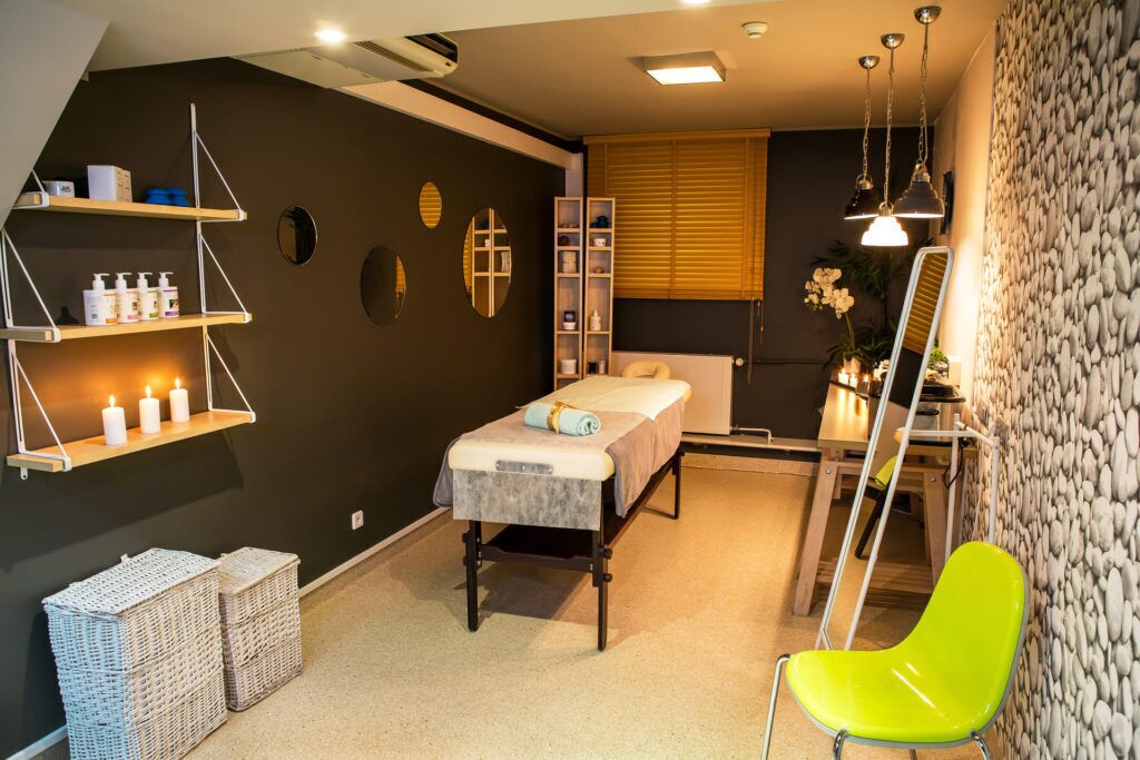 Una habitación con una cama, una camilla de masaje, estantes con artículos de aseo, un espejo y una silla luminosa.