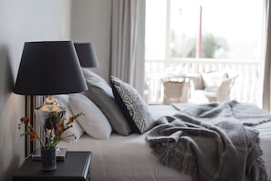 Un lit bien fait, recouvert d’une couverture grise, une lampe de chevet et un vase rempli de fleurs.