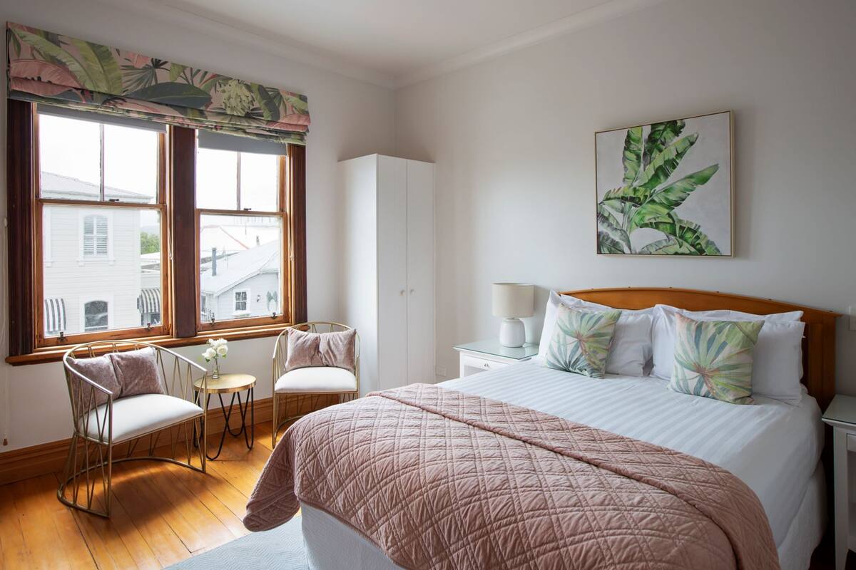 A bedroom with a large bed, a chair, a small table, and a window with a view of buildings.