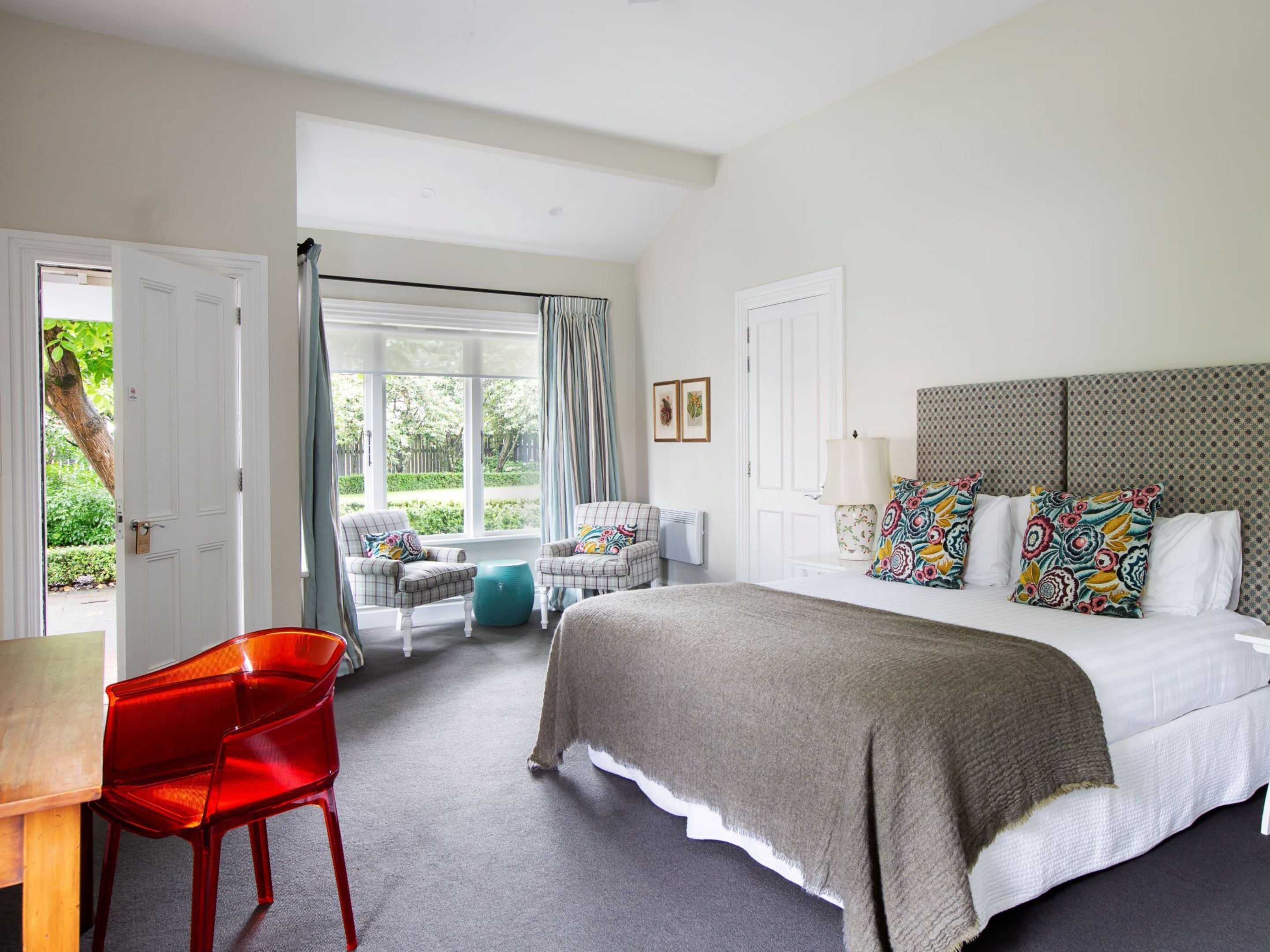A bedroom with a bed, two armchairs, a red chair, a window with curtains, and a view of greenery outside.