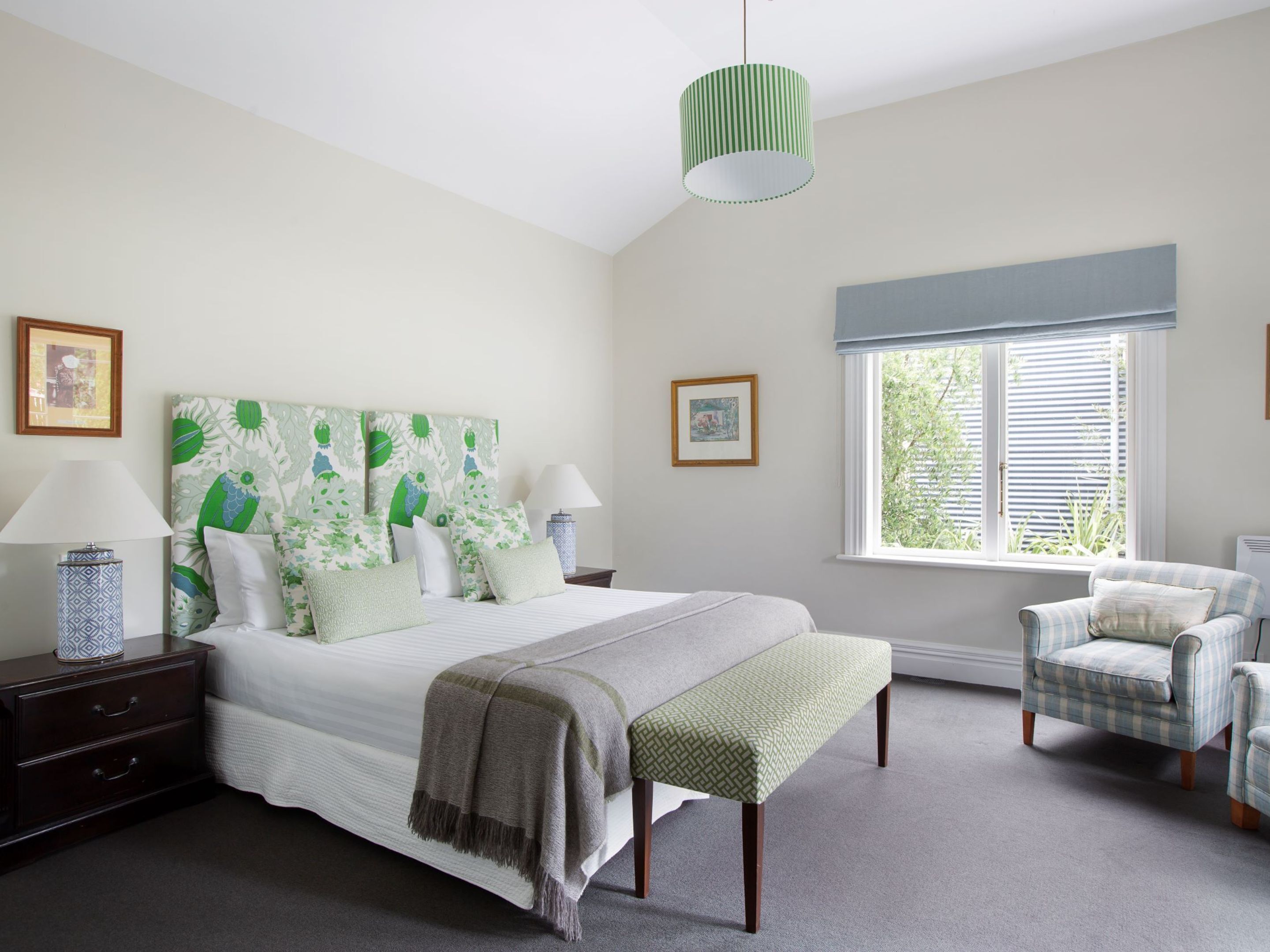 A bedroom with a large bed, a bench, a chair, a nightstand, a lamp, and a window with blinds.