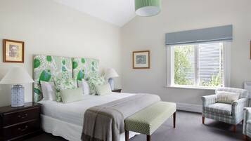 A bedroom with a large bed, a bench, a chair, a nightstand, a lamp, and a window with blinds.